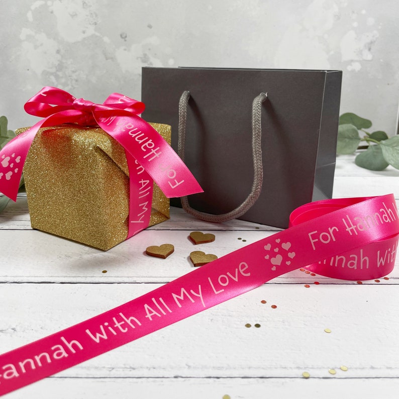 May include: A pink satin ribbon with the text "For Hannah with All My Love" printed in white. The ribbon is wrapped around a gold glitter gift box and a gray gift bag.
