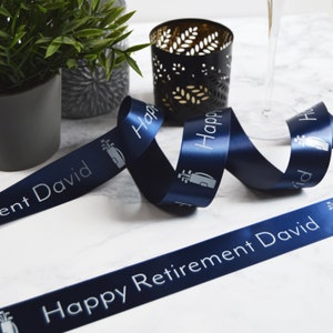 Pode incluir: Fita de cetim azul marinho com o texto "Happy Retirement David" e um gráfico de saco de golfe. A fita está enrolada sobre uma superfície de mármore branco. Um castiçal preto e uma pequena planta em vaso estão no fundo.
