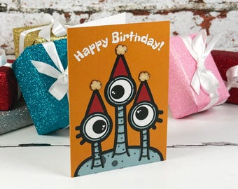 Könnte beinhalten: Eine orangefarbene Geburtstagskarte mit drei einäugigen Cartoon-Kreaturen, die rote Partyhüte mit beigen Bommeln tragen. Der Text Happy Birthday ist in weißer Schrift oben auf der Karte über den abgebildeten Kreaturen aufgedruckt.
