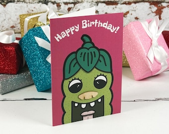 Könnte beinhalten: Eine magentafarbene Grußkarte mit dem Text Happy Birthday in weißen Buchstaben. Die Karte zeigt eine Cartoon-Illustration einer grünen, texturierten, gurkenartigen Figur mit großen Augen, einer beigen Nase und einem offenen Mund mit Zähnen.