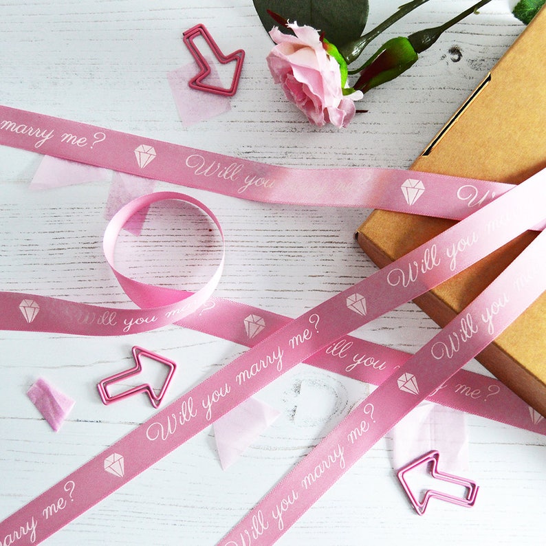 May include: Pink satin ribbon with the text "Will you marry me?" printed in white. The ribbon is decorated with small diamond shapes.