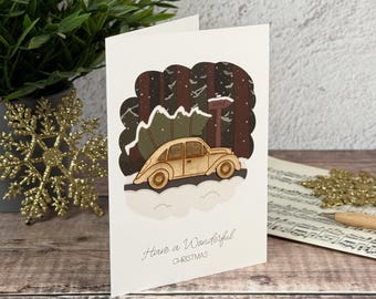 Könnte beinhalten: Eine weiße Grußkarte mit einer mehrschichtigen Illustration eines beigen Autos, das einen grünen Weihnachtsbaum auf dem Dach durch einen dunklen Wald transportiert. Ein Wegweiser zeigt HOME. Der Text Have a Wonderful CHRISTMAS steht unten in Grau.