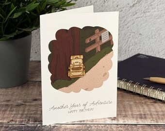 Könnte beinhalten: Eine weiße Grußkarte mit einer zentralen Illustration eines hellbraunen Wanderrucksacks, der auf einem Feldweg neben einem großen Baum und einem Holzzaun steht. Der Text lautet Another Year of Adventure und HAPPY BIRTHDAY in Schwarz.