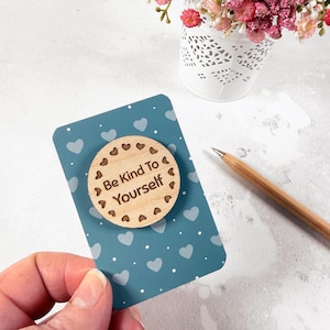 Può includere: Un magnete circolare in legno con la scritta "Be Kind To Yourself" su una carta blu con cuori bianchi. Il magnete è circondato da piccole forme di cuore. Una matita e un piccolo vaso di fiori bianco sono sullo sfondo.