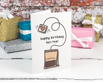 Könnte beinhalten: Eine weiße Grußkarte mit einer Cartoon-Illustration eines braunen Teebeutels, dessen Schnur mit einem Etikett mit einem rosa Herz verbunden ist. Der Text auf der Karte lautet "happy birthday bes-tea!" in schwarzen Kleinbuchstaben.