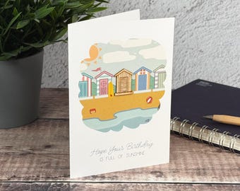 Könnte beinhalten: Eine weiße Grußkarte mit einer farbenfrohen Illustration von Strandhäusern an einem sandigen Ufer mit einer Sonne am Himmel. Der Text auf der Karte lautet, Hope Your Birthday IS FULL OF SUNSHINE. Die Karte steht aufrecht auf einer Holzoberfläche.