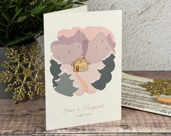 Könnte beinhalten: Eine weiße Grußkarte mit einer zentralen Illustration einer Holzhütte in einer verschneiten Berglandschaft in Rosa- und Grautönen. Der Text Have a Magical CHRISTMAS ist unten in Grau gedruckt. Ein kleiner Holzhütten-Ausschnitt ist befestigt.