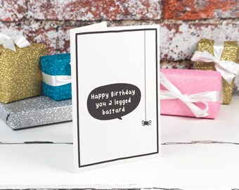 Könnte beinhalten: Eine weiße Grußkarte mit schwarzem Rand, die eine schwarze Sprechblase mit dem Text Happy Birthday you 2 legged bastard und eine kleine schwarze Spinne an einem vertikalen Faden auf der rechten Seite der Karte zeigt.