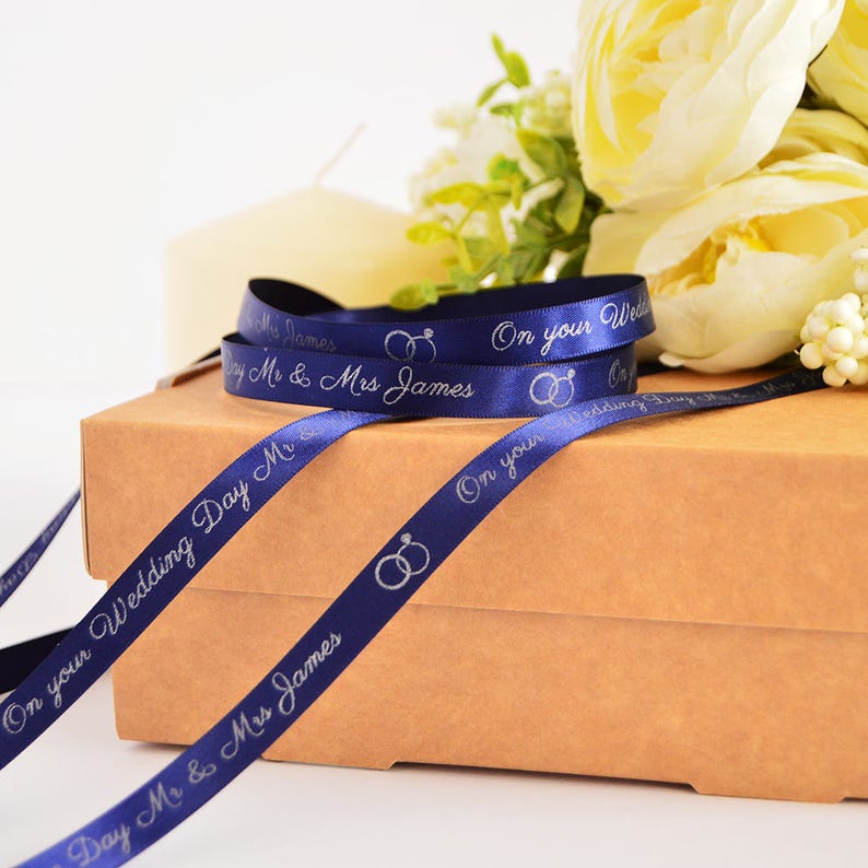 K&ouml;nnte beinhalten: Ein dunkelblaues Satinband mit silbernem Foliendruck, der "On your Wedding Day" und "Mr & Mrs James" sowie zwei ineinander verschlungene Eheringe zeigt. Das Band ist aufgerollt und liegt auf einer braunen Pappschachtel.