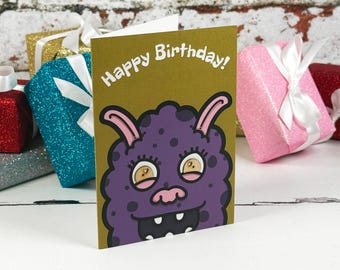 Könnte beinhalten: Eine Grußkarte mit goldenem Hintergrund, einer lila Monsterillustration und dem weißen Text Happy Birthday. Das Monster hat zwei rosa Ohren, große Augen und einen breiten Mund mit sichtbaren Zähnen. Die Karte ist von glitzernden Geschenkboxen umgeben.