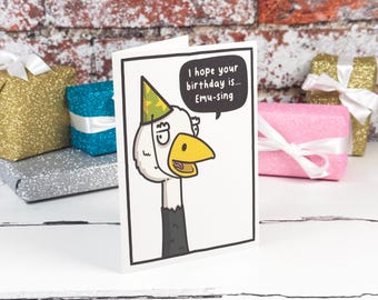 Könnte beinhalten: Eine weiße Grußkarte mit einem Cartoon-Emu, der einen grün-gelb gemusterten Partyhut trägt. Eine schwarze Sprechblase enthält den Text, I hope your birthday is... Emu-sing. Die Karte hat einen schwarzen Rand und steht auf einer weißen Fläche.