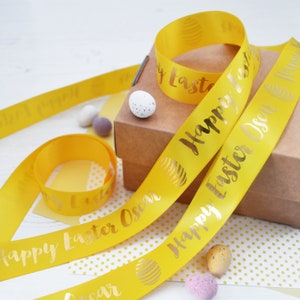 May include: Yellow satin ribbon with gold foil lettering that reads "Happy Easter Oscar". The ribbon is wrapped around a brown gift box and there are small chocolate eggs scattered around.