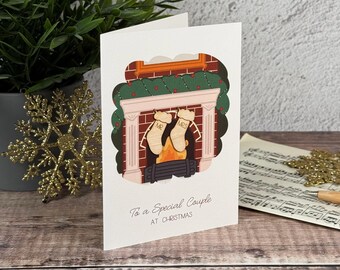 Könnte beinhalten: Eine weiße Grußkarte mit einer Kaminillustration, an deren Kaminsims zwei Holzstrümpfe mit der Aufschrift MR und MRS hängen. Der Text lautet To a Special Couple AT CHRISTMAS. Die Karte ist mit einer grünen Girlande und Feuer verziert.
