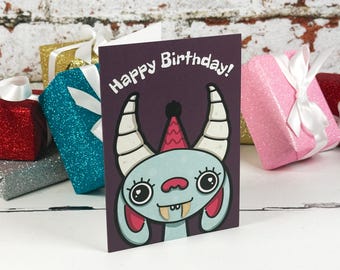 Könnte beinhalten: Eine dunkelviolette Geburtstagskarte mit einer Cartoon-Illustration eines hellblauen Monsters mit großen Augen, zwei Hörnern und einem rosa Partyhut. Der Text Happy Birthday ist in Weiß oben auf der Karte über der Monstergrafik aufgedruckt.