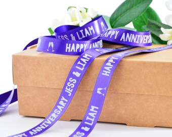 May include: A purple satin ribbon featuring white printed text that reads "HAPPY ANNIVERSARY" and "JESS & LIAM" along with a graphic of two champagne flutes. The ribbon is draped over a rectangular brown cardboard box with white flowers nearby.