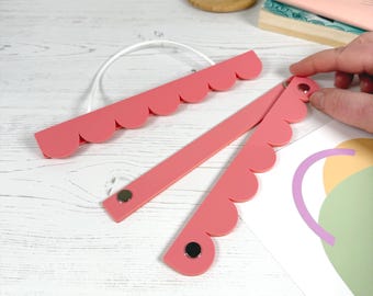 Könnte beinhalten: Ein Set aus zwei korallenfarbenen magnetischen Plakatleisten mit gewelltem Rand. Jede Leiste besteht aus zwei flachen Streifen, die durch kleine silberfarbene Magnete verbunden sind, und einer weißen Schnur am oberen Teil zum Aufhängen.