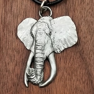 May include: A silver-colored elephant pendant with detailed features, including tusks and textured skin, hangs from a black cord necklace. The pendant is suspended by a silver ring. The background is a wooden surface.