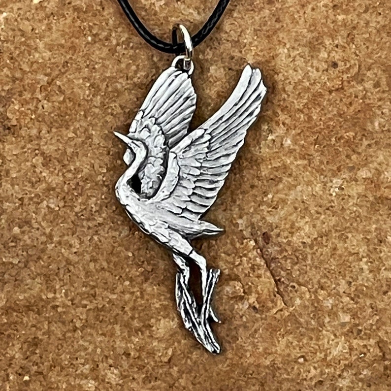 May include: A silver pendant in the shape of a crane with its wings spread. The crane is in flight and has a long neck and legs. The pendant is on a black cord.