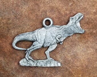 May include: Silver-toned pewter pendant featuring a detailed sculpture of a Tyrannosaurus Rex dinosaur with its mouth open. The pendant has a loop at the top for attaching a chain.