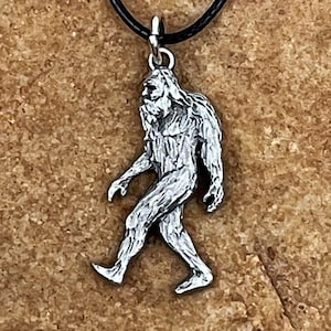May include: A silver pendant necklace featuring a detailed depiction of Bigfoot walking. The pendant is attached to a black cord.