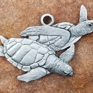 May include: A silver charm featuring two sea turtles, one on top of the other, with a loop for attaching to a necklace or bracelet.