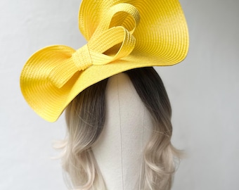 May include: A bright yellow sun hat with a large bow detail. The hat is made of a woven material and has a wide brim. The hat is displayed on a mannequin head with blonde and brown hair. The hat is a stylish accessory.