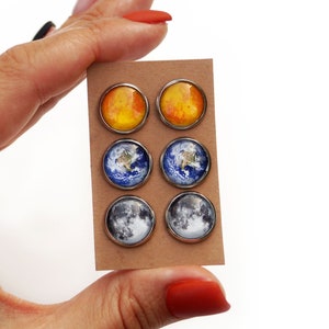 May include: Six pairs of stud earrings featuring images of the sun, earth, and moon. The earrings are displayed on a brown card.