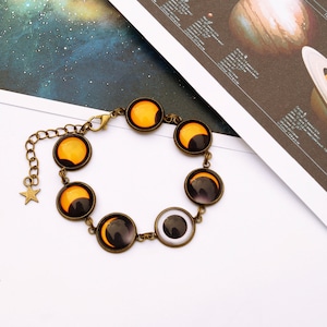 May include: A gold-toned bracelet with a chain and seven round charms. The charms feature images of the moon in different phases, including a full moon, a crescent moon, and a new moon. The bracelet is on a white surface.
