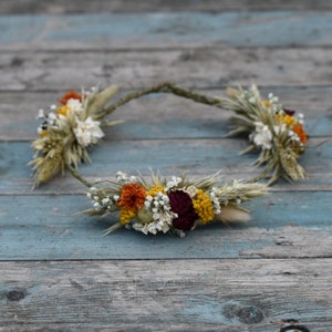 May include: A dried flower crown with a rustic, natural look. The crown is made with dried wheat, white flowers, red flowers, and yellow flowers.