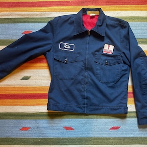 May include: A navy blue work jacket with a red interior lining, a zip-up front, and two chest pockets. The jacket features a name patch that says "Rita" and a Pontiac logo patch.