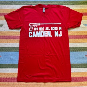 Può includere: Maglietta rossa con una grafica bianca di una pistola e il testo "It's not all good in Camden, NJ".