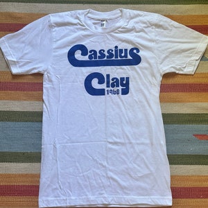 May include: White t-shirt with a blue graphic print of "Cassius Clay 1960" in a retro font.