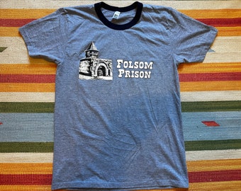 May include: Gray ringer t-shirt with a navy blue collar and sleeve trim. The shirt features a black and white graphic of a building and the words "FOLSOM PRISON" in white block letters.