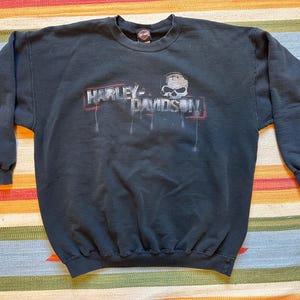 May include: Black Harley-Davidson sweatshirt with a graphic featuring the brand's logo and a skull. The logo is in red and white, with a distressed look. The sweatshirt has long sleeves and a crew neck.