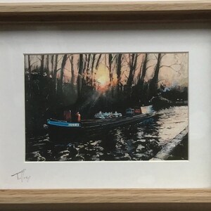 May include: A framed print featuring a dark barge on a river at sunset. The scene depicts a long boat with a blue bow, silhouetted trees, and a bright sun glowing through the branches. The artwork is enclosed in a light-coloured wooden frame.