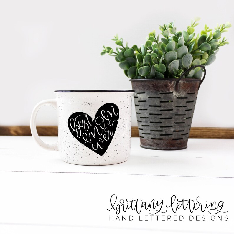 Może przedstawiać: Biały, nakrapiany ceramiczny kubek z czarną obw&oacute;dką i uchwytem. Czarny serce z napisem "best mom ever" znajduje się na kubku. Mały metalowy kubełek z zielenią stoi po prawej stronie kubka.
