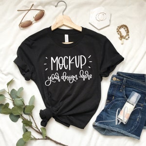 Puede incluir: Camiseta negra con el texto "MOCKUP your design here" en escritura blanca. La camiseta se muestra en una percha de madera. También se ven gafas de sol, joyas y pantalones cortos vaqueros.