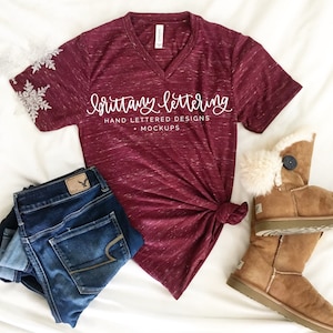 Puede incluir: Una camiseta burdeos con cuello en V con el texto "brittany lettering" y "hand lettered designs + mockups". La camiseta está combinada con vaqueros azules y botas de invierno marrones.