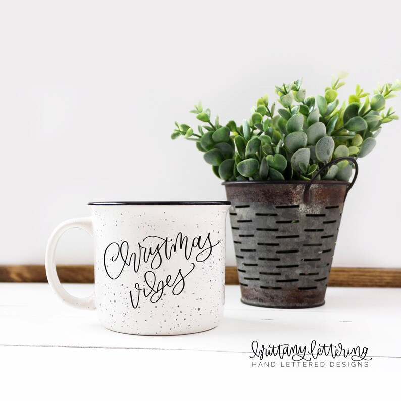 May include: White speckled ceramic mug with black rim and handle. The mug features the words "Christmas vibes" in elegant black script. Beside the mug is a small metal bucket filled with green foliage. The words "Brittany Lettering" are at the bottom.
