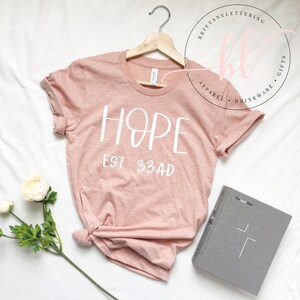 May include: A pale pink t-shirt with the word "HOPE" in white script and "EST. 33 AD" printed below. The shirt is displayed on a wooden hanger. A grey book with a white cross and a white flower are also visible.