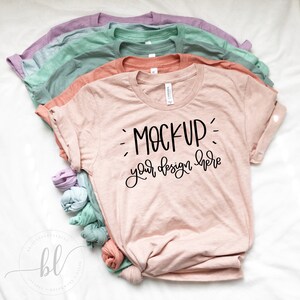 Puede incluir: Una pila de cinco camisetas en colores pastel: rosa claro, coral, verde menta, verde claro y lavanda. La camiseta delantera tiene la frase "MOCKUP your design here" en negro. Las camisetas están dobladas sobre una superficie blanca.
