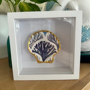May include: A white square shadow box frame containing a single scallop shell. The shell features a white interior with dark blue painted coral-like patterns and a metallic gold painted edge, mounted against a plain white background.