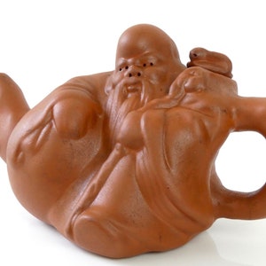 Puede incluir: Una tetera de terracota con forma de figura sentada con una larga barba y una pequeña figura en su hombro. La tetera tiene un pico y una asa.
