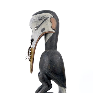 May include: A wooden carving of a bird with a long beak, painted in black, white and brown. The bird is standing on a round base and has a long, thin beak with a small, white object in its mouth.