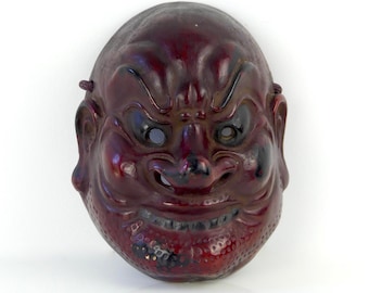 May include: A red wooden mask with a fierce expression. The mask has black eyes and a wide, open mouth with sharp teeth. The mask is carved with intricate details and has a textured surface.