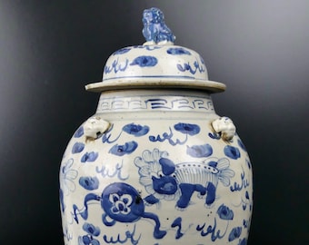 May include: A large, white porcelain jar with a blue and white design. The jar has a lid with a lion finial and is decorated with stylized lions and floral motifs. The jar is approximately 18 inches tall.