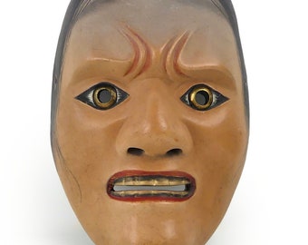 May include: A traditional Japanese Noh mask with a carved wooden face, painted in shades of brown and red. The mask has exaggerated features, including a wide mouth, large eyes, and a furrowed brow. The eyes are painted with gold circles, and the mouth is open in a grimace.