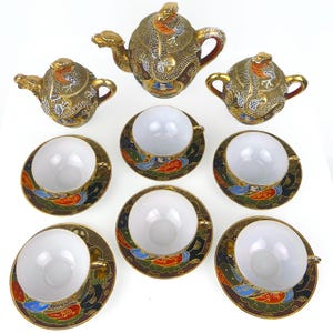 May include: A tea set featuring a teapot, a lidded sugar bowl, a lidded creamer, and six teacups with saucers. Each piece is decorated with a raised gold dragon motif on a dark background, accented with red, blue, and green enamel details.