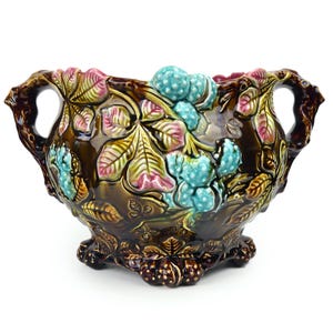 Puede incluir: Un tazón grande de cerámica marrón con dos asas y tres patas. El tazón está decorado con hojas y bayas de colores en relieve.