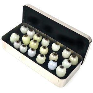 May include: A rectangular silver-toned metal storage tin containing a collection of eighteen small, spherical glass eye cabochons. Each piece features a smooth, off-white sclera and a centered, dark brown iris, arranged neatly on a black interior base.